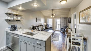 A modern kitchen with a marble countertop, grey cabinets, and a built-in dishwasher. There is a dining area with a table and chairs, and a living room with a sofa and a ceiling fan.