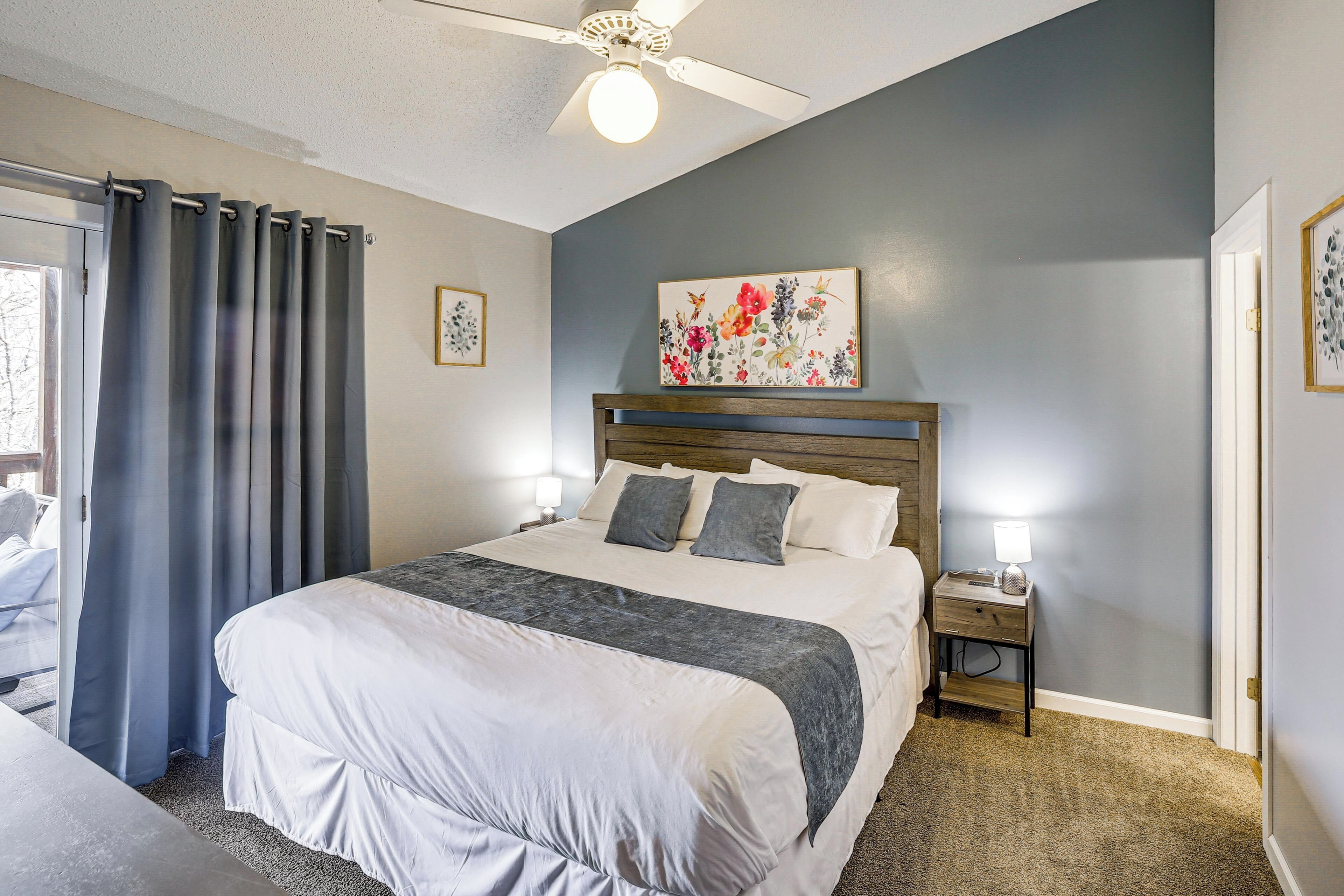A bedroom with a large bed, a ceiling fan, a nightstand with a lamp, and a floral painting on the wall.