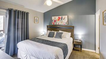 A bedroom with a large bed, a ceiling fan, a nightstand with a lamp, and a floral painting on the wall.