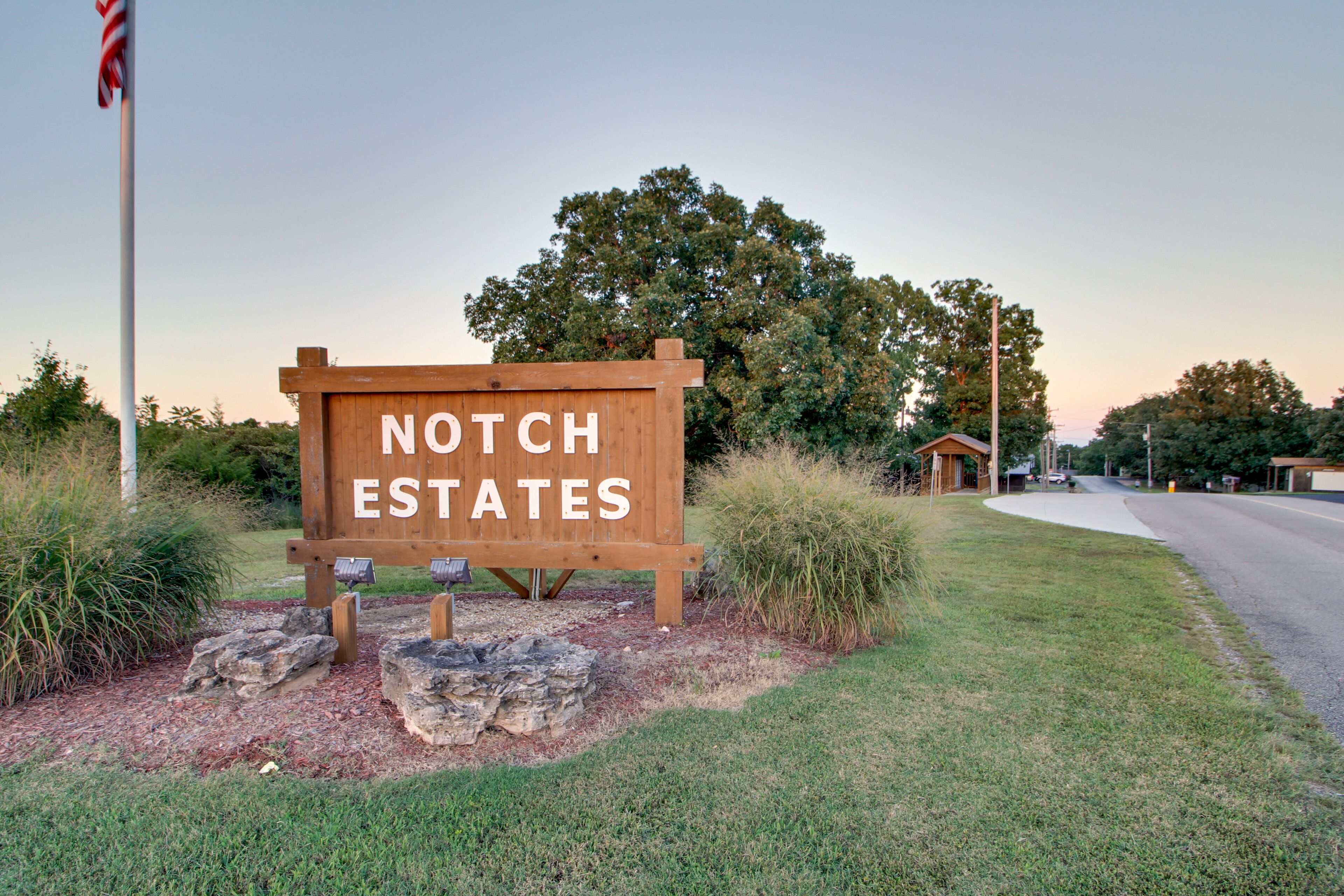 A wooden sign with "NOTCH ESTATES" written on it, surrounded by grass and rocks, with a road and a building in the background.
