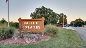 A wooden sign with "NOTCH ESTATES" written on it, surrounded by grass and rocks, with a road and a building in the background.