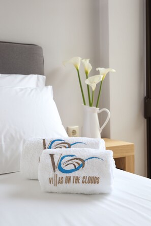 A neatly made bed with white linens, a white vase with flowers, and a towel with a logo and text. - Villas On The Clouds (Lefkada)
