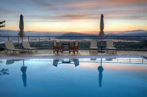 An infinity pool with a view of a sunset over mountains, surrounded by lounge chairs and umbrellas. - Villas On The Clouds (Lefkada)