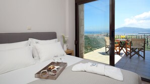 Hotelkamer met een bed, een dienblad met ontbijtartikelen, een raam met uitzicht op zee en bergen, en een balkon met een tafel en stoelen.