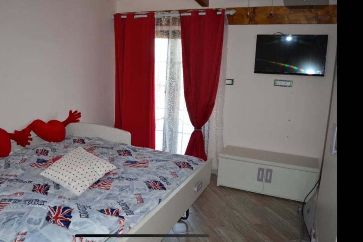 A bedroom with a bed featuring a patterned bedspread, a white bedside table, a red curtain, and a television mounted on the wall.