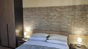 A bedroom with a stone wall headboard, a bed with white linens, and two bedside lamps. - Villa Carmen (Mondragone)