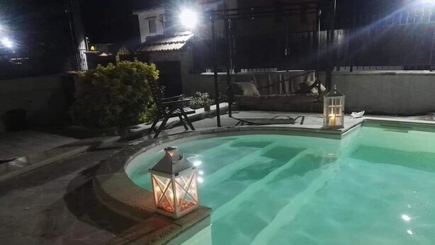 A nighttime swimming pool with a curved edge, surrounded by a patio area with outdoor furniture and two lit lanterns. - Villa Carmen (Mondragone)
