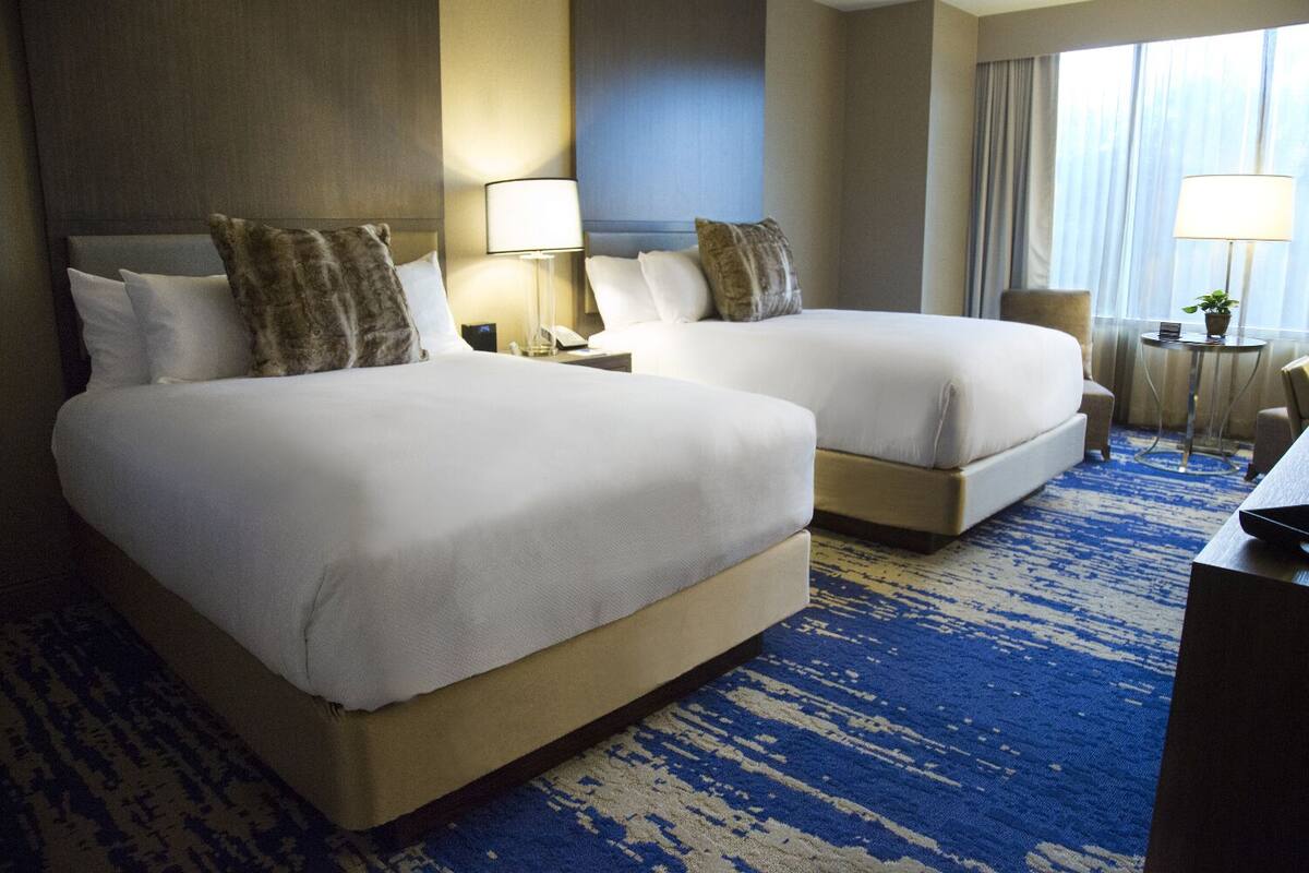 A hotel room with two beds, a desk, a chair, a lamp, and a view of the outdoors.