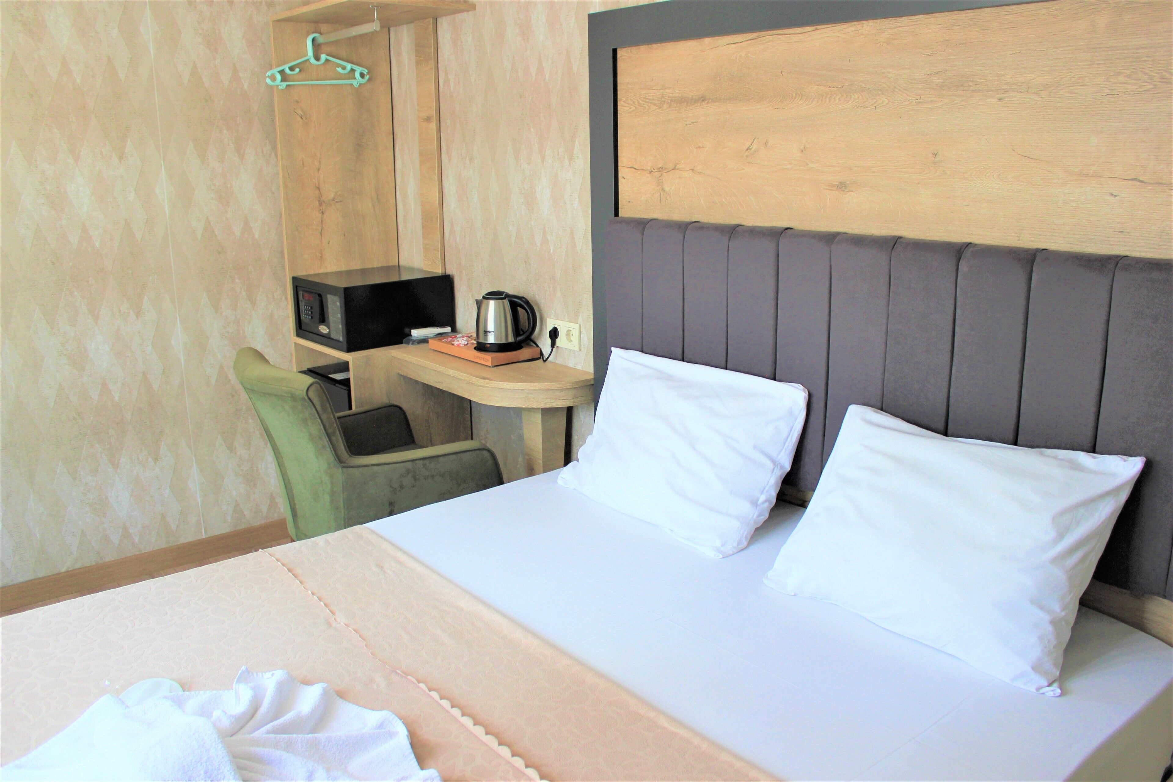 A hotel room with a bed, a small desk with a kettle, and a chair.