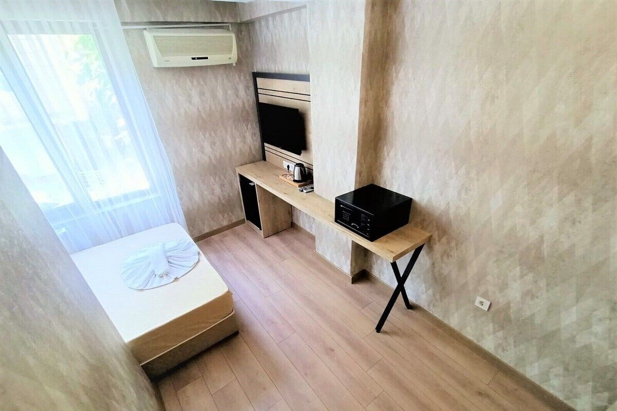 A compact room with a bed, a small desk, a chair, and a window with sheer curtains.