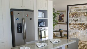 A modern kitchen with white cabinetry, a stainless steel refrigerator, a built-in oven, and a granite countertop. There are two white chairs and a small table with a decorative plant in the background. - Penelope Beach House (San Andrés)