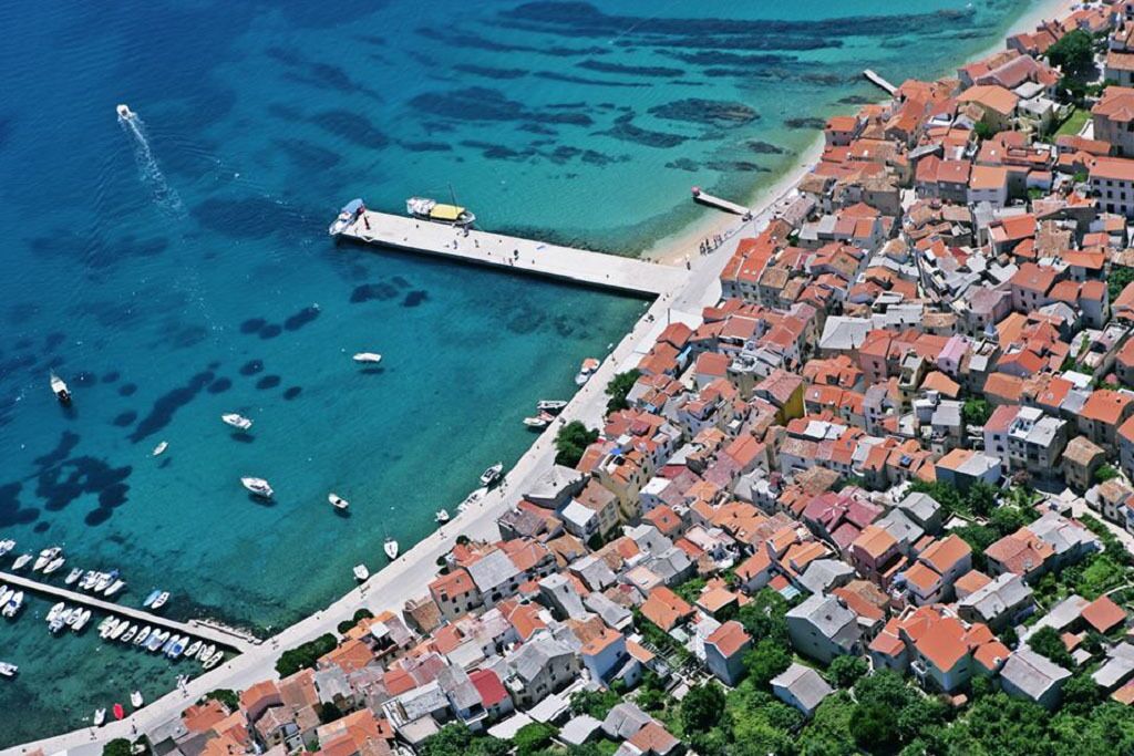An aerial view of a coastal town with a pier extending into the sea, surrounded by densely packed buildings with red-tiled roofs.