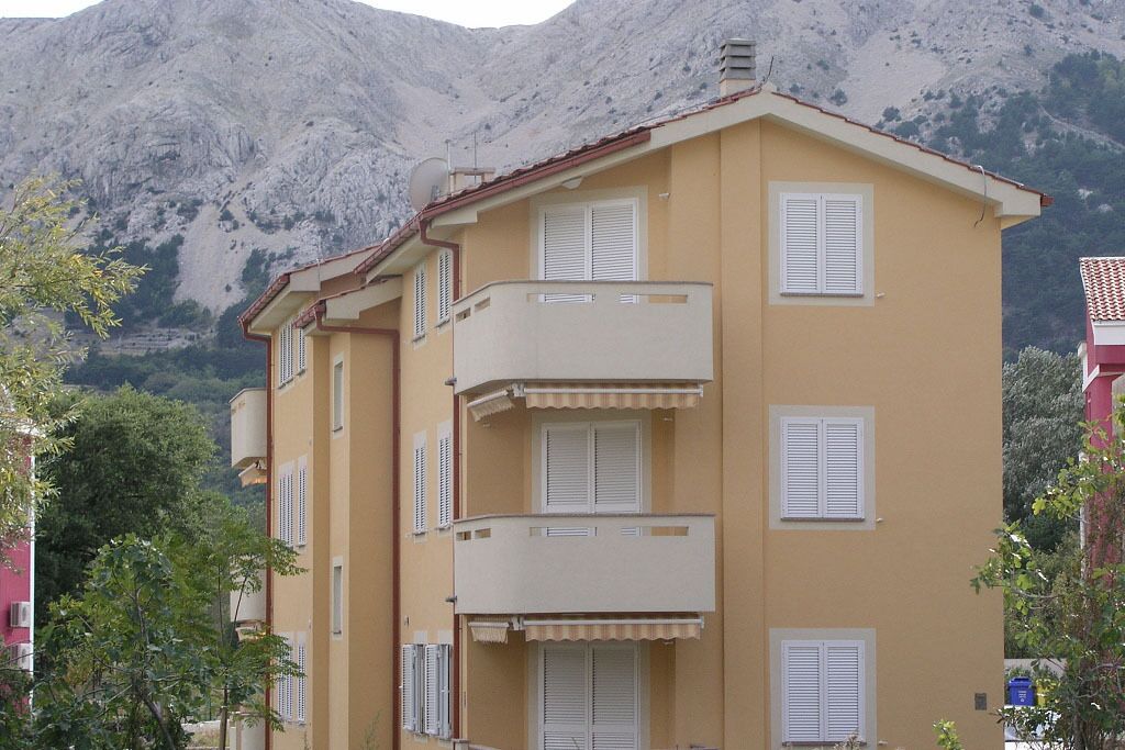 A multi-story building with balconies and white shutters, set against a mountainous backdrop.