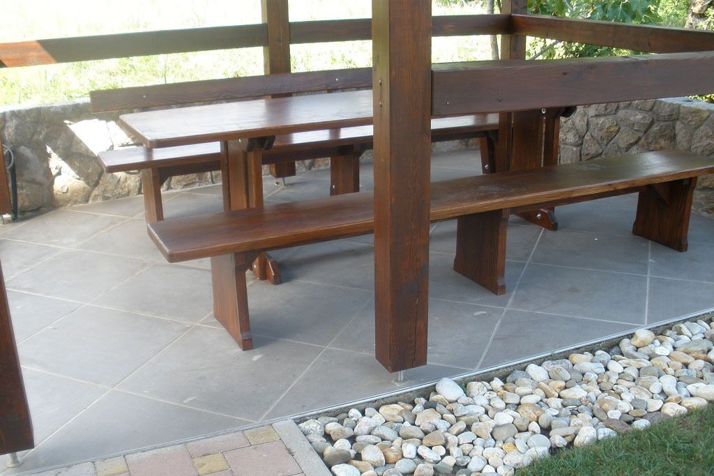 A wooden picnic table with benches, set on a stone-tiled patio with a pebble border.