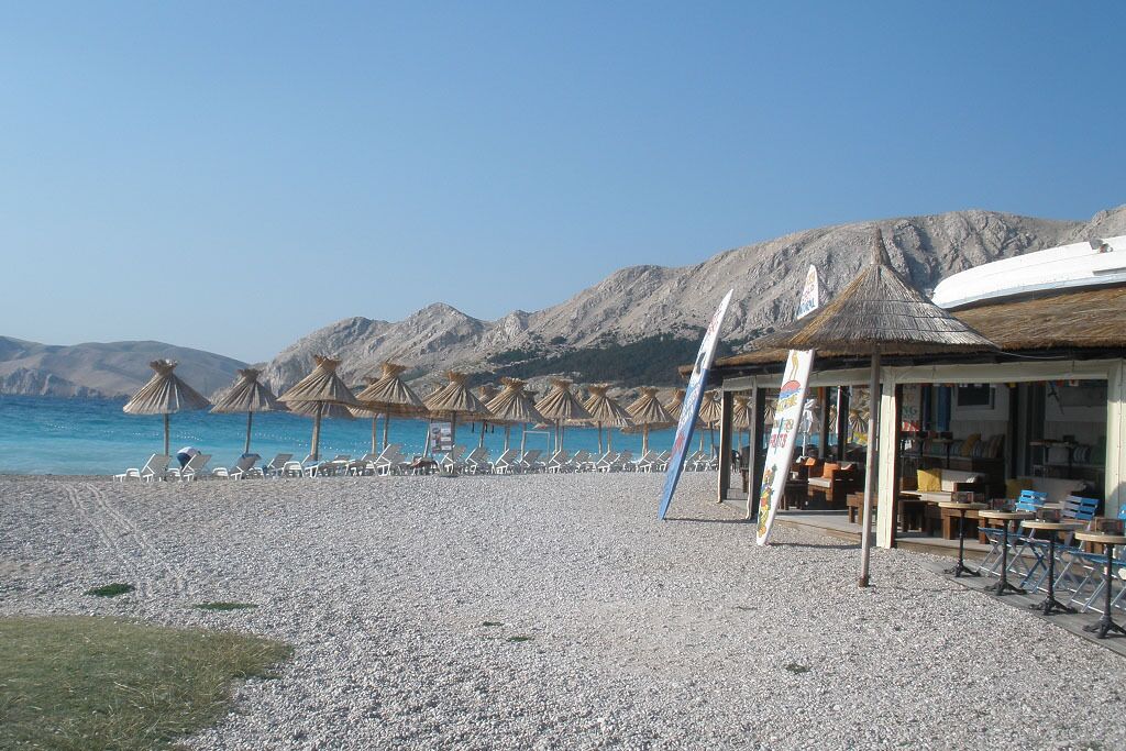 A beachside restaurant with outdoor seating, umbrellas, and a view of the sea and mountains.