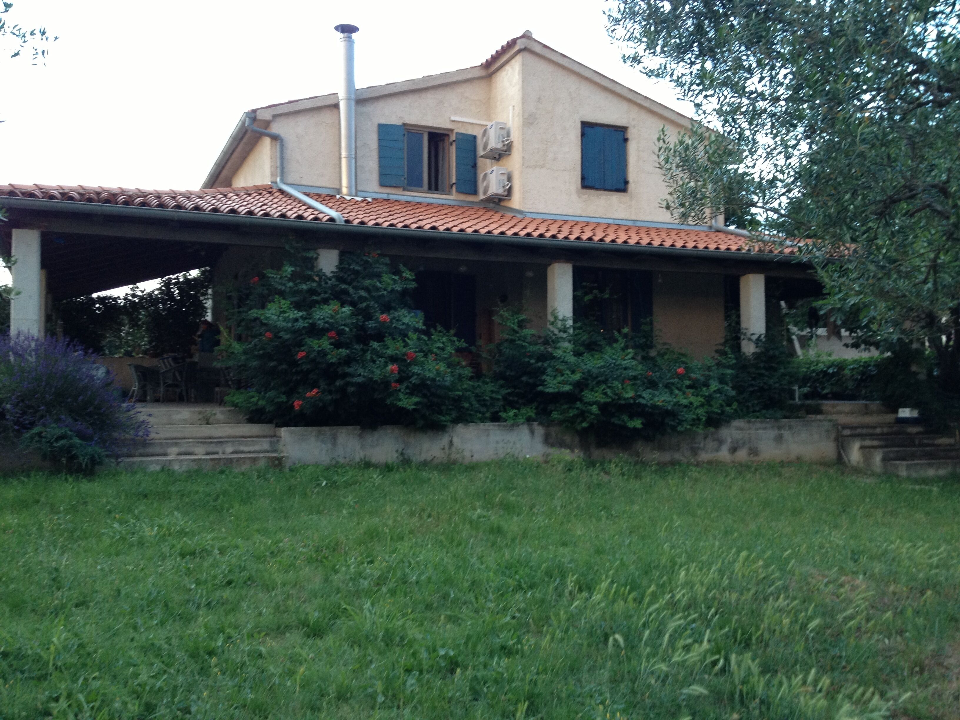 A two-story house with a tiled roof, a covered porch, and a garden with various shrubs.