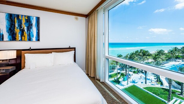 Habitación de hotel con una cama grande, una mesita de noche, un cuadro y vistas a la playa y al océano.