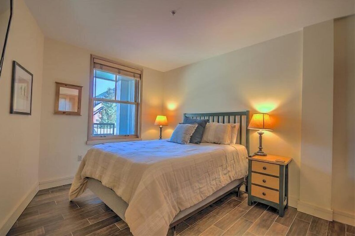 A bedroom with a bed, two bedside lamps, a nightstand, a window, and a framed picture on the wall.