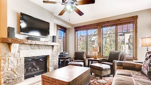 A living room with a stone fireplace, leather furniture, a flat-screen TV, and large windows. - Luxury 3 Bedroom Breckenridge Vacation Rental with Mountain Views Steps from Historic Main Street (Breckenridge)