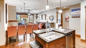 A modern kitchen with a central island, wooden cabinets, and a built-in dishwasher. There are two pendant lights, a large window, and a dining area with orange chairs. - Luxury 3 Bedroom Breckenridge Vacation Rental with Mountain Views Steps from Historic Main Street (Breckenridge)