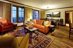 A cozy living room with a red armchair, a brown sofa, and a coffee table. There is a dining table with chairs and a painting on the wall.