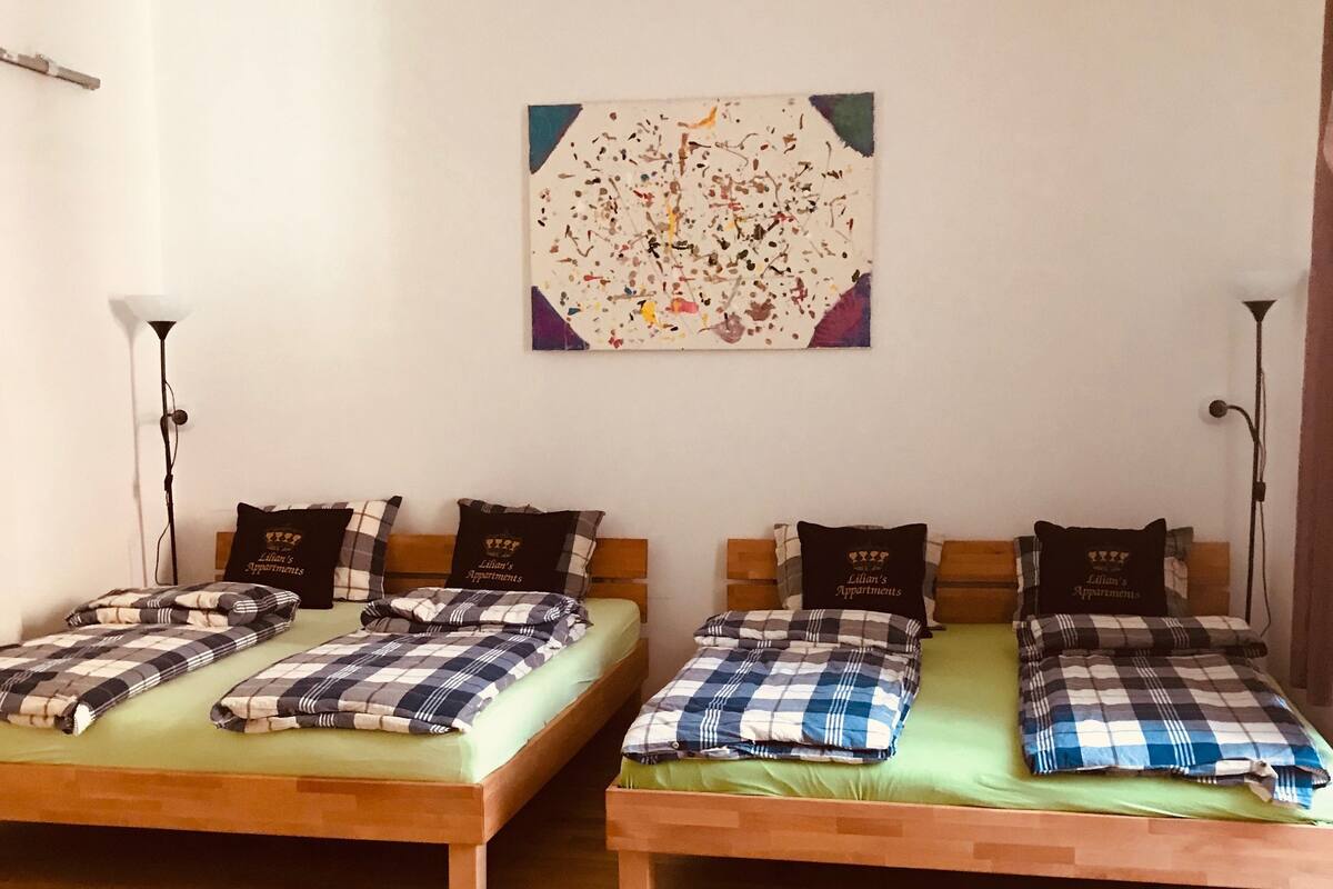 A room with two wooden beds, each with a green mattress and checkered bedding. There is a colorful abstract painting on the wall above the beds.