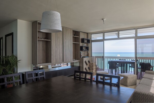 A modern living room with a large dining table, built-in wooden shelving, and a view of the ocean.