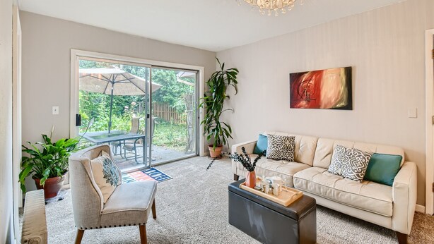 Uma sala de estar espaçosa com um sofá bege, uma mesa de centro e um lustre. Há uma porta de vidro deslizante que dá para um pátio externo com uma mesa e cadeiras.