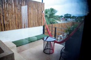 Un balcón con pared de bambú, una hamaca roja, una mesita redonda y un banco verde con cojín.