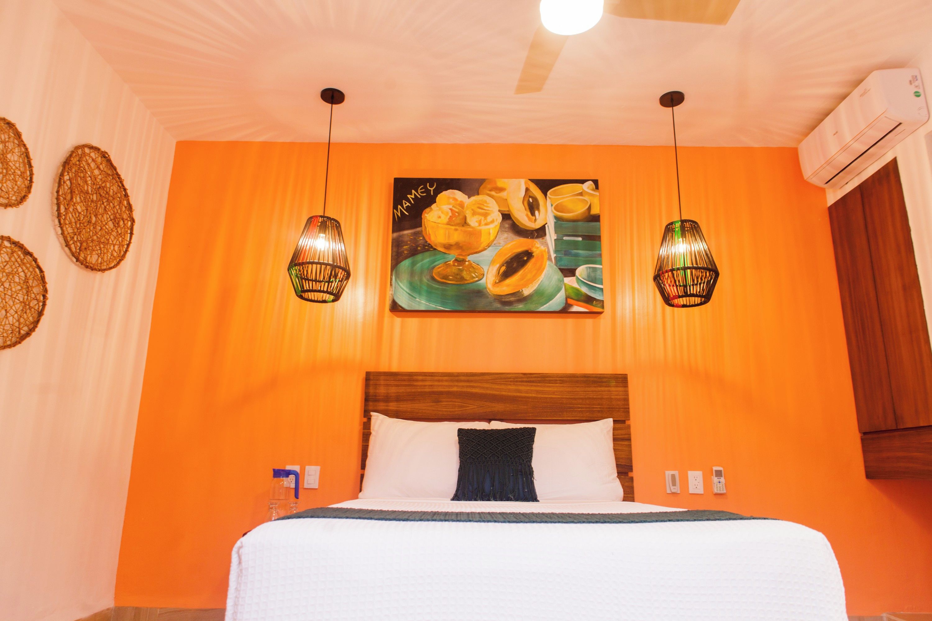 Habitación de hotel con pared naranja, cama con ropa de cama blanca, dos lámparas colgantes y un cuadro sobre la cama.