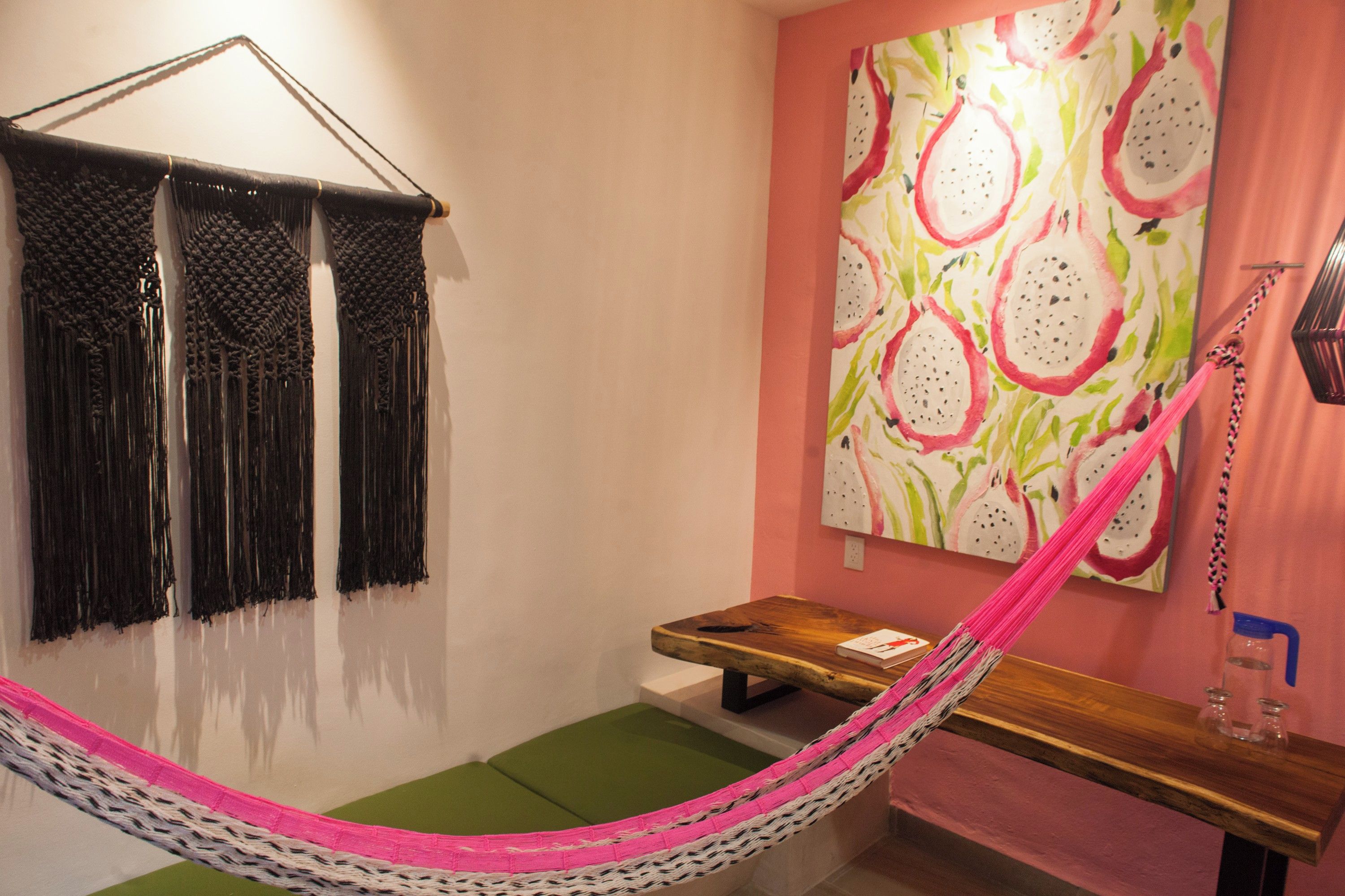 A hammock chair with a pink and white woven seat, a wooden bench, a small table with a book, and a wall with a colorful abstract painting.