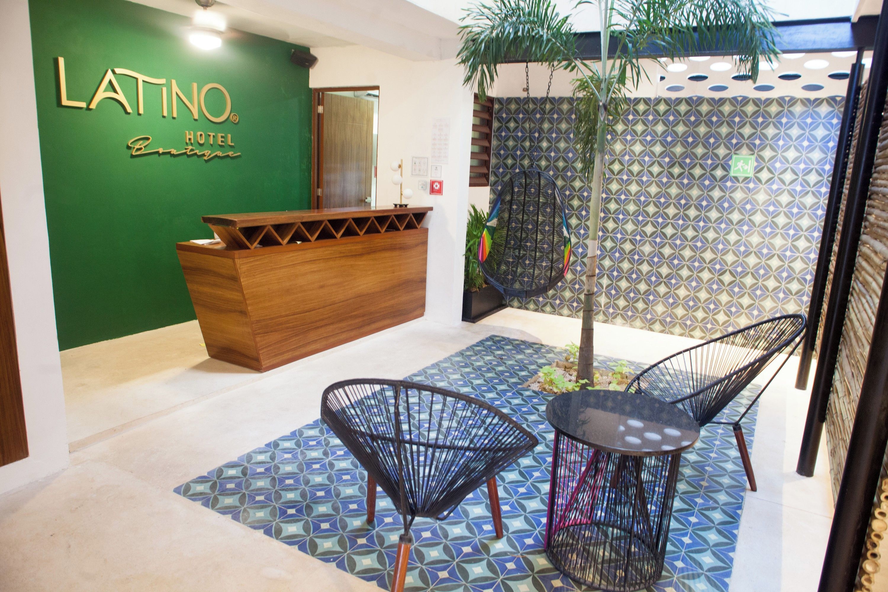 A hotel reception area with a wooden desk, two black chairs, patterned floor tiles, and a green wall with the hotel's name.