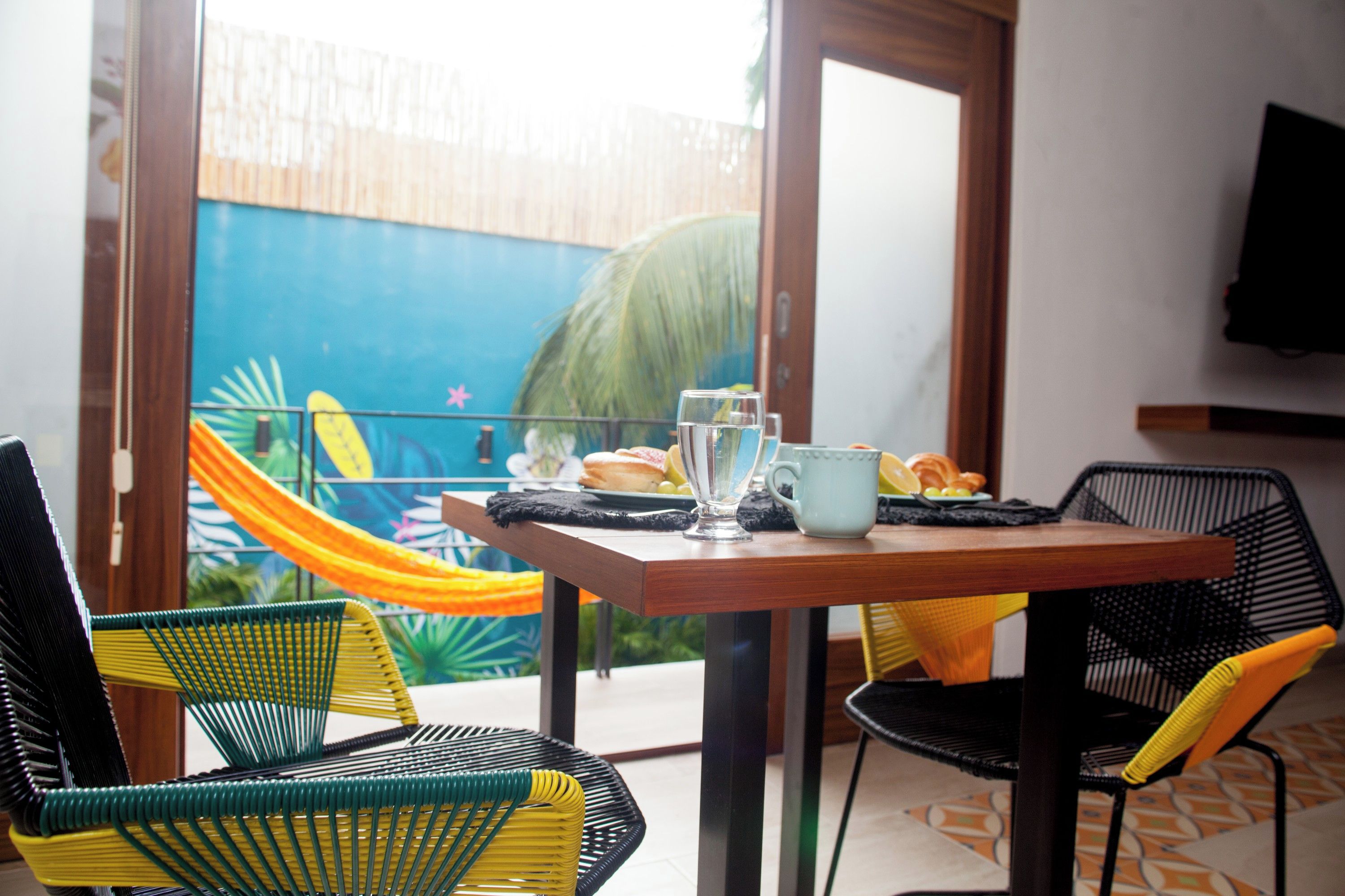 Un comedor con una mesa dispuesta para dos personas, sillas y vista al cielo azul y palmeras a través de un ventanal.