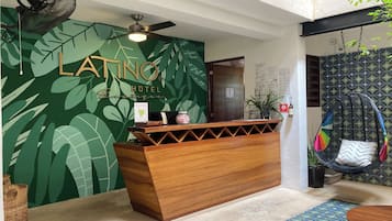 Un mostrador de recepción con detalles de madera, un ventilador de techo en lo alto y una pared con motivos de hojas tropicales.