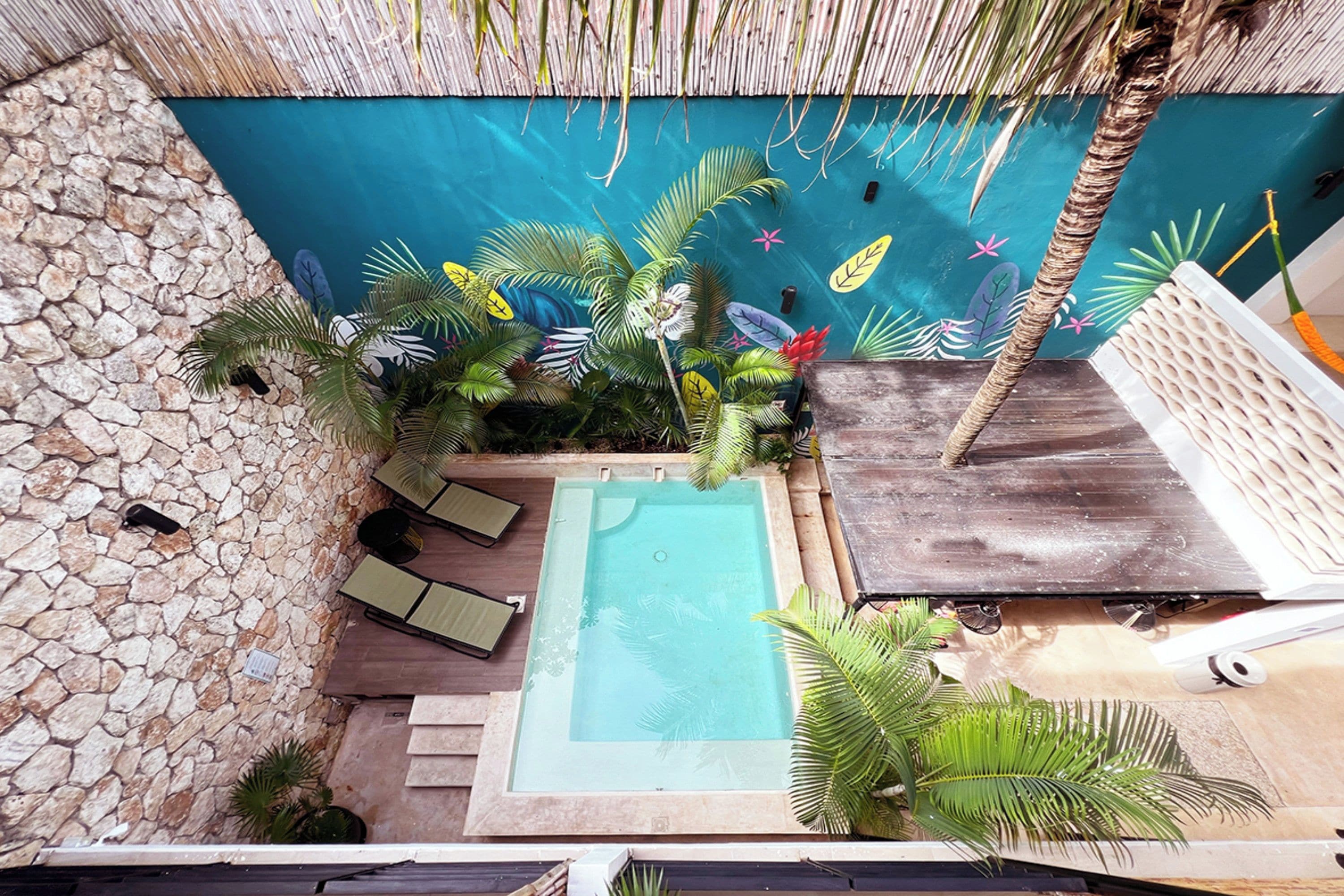 A rooftop pool area with a stone wall, lounge chairs, and tropical plants.