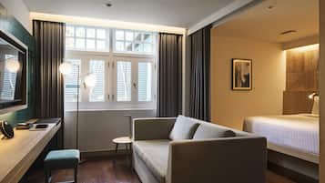 Una habitación de hotel con sofá, una mesita, una mesita de noche, una cama, una ventana con cortinas y un cuadro en la pared.