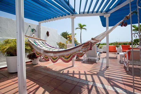 Un hamac sous une auvent bleue, des murs blancs et des chaises orange sur une terrasse carrelée.