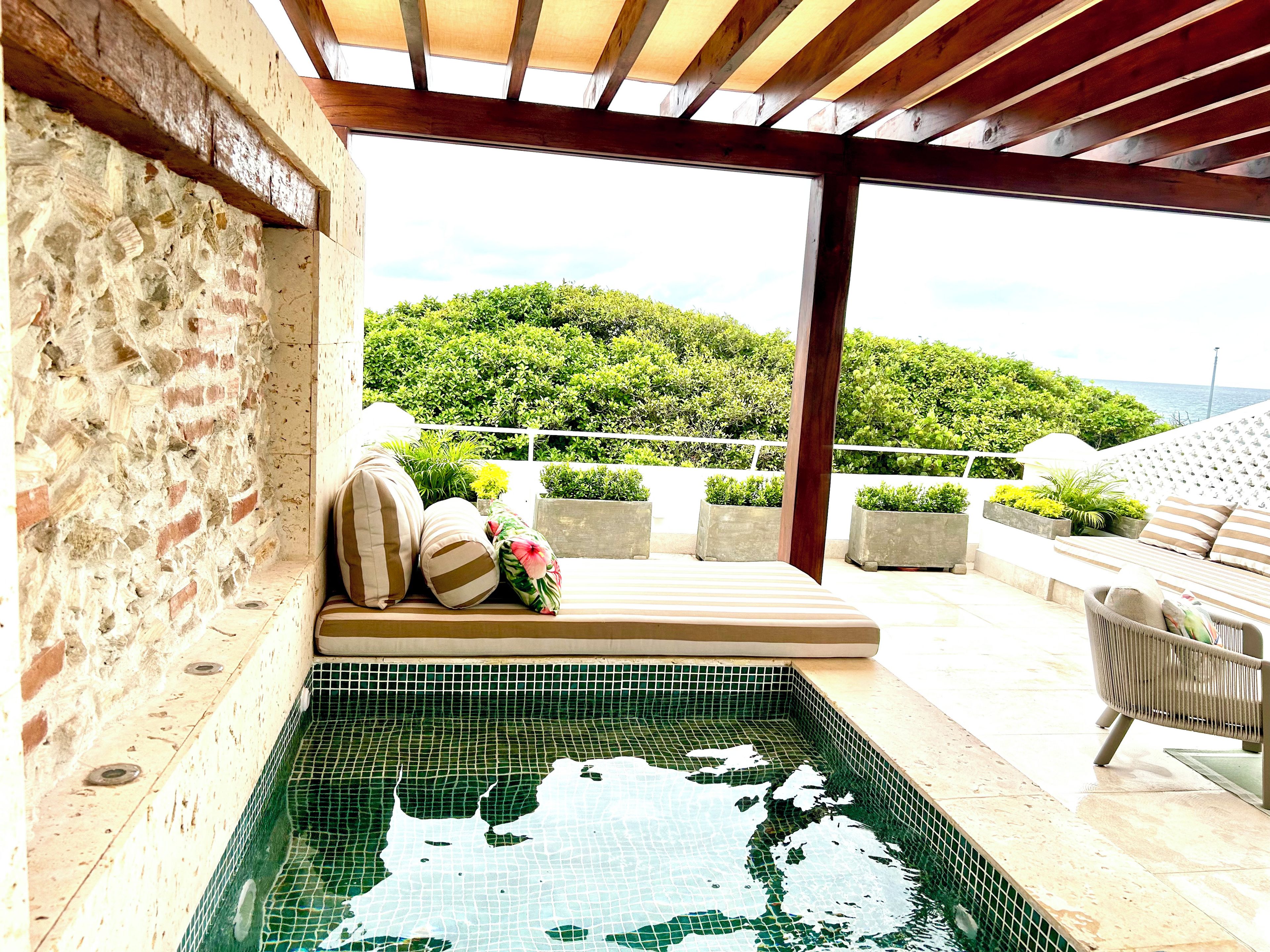 A pool area with a tiled floor, a bench with cushions, and a view of a green landscape.
