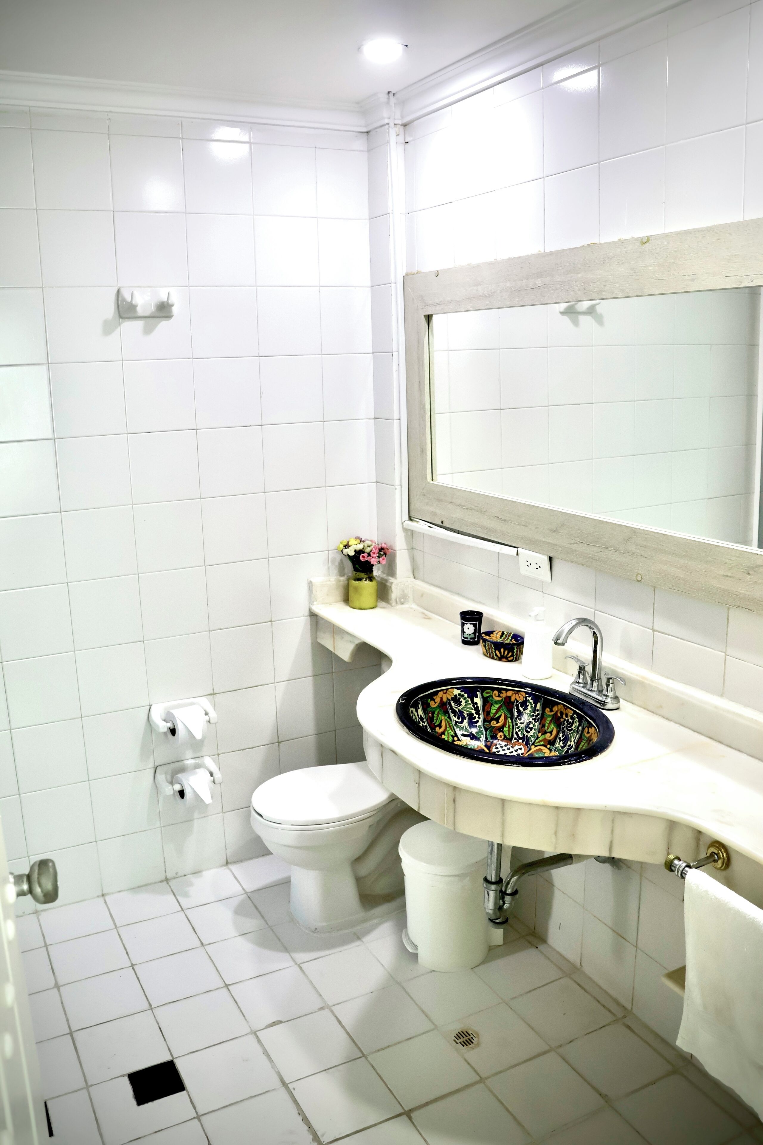 Un baño con paredes de azulejos blancos, un inodoro blanco, un lavabo con una pileta de colores y un espejo sobre el lavabo.