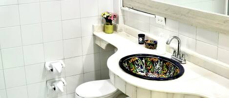 Un baño con paredes de azulejos blancos, un inodoro blanco, un lavabo con una pileta de colores y un espejo sobre el lavabo.