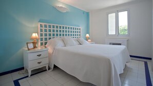 A bedroom with a white bed, two bedside tables, a white nightstand, a window, and a wall-mounted air conditioner.