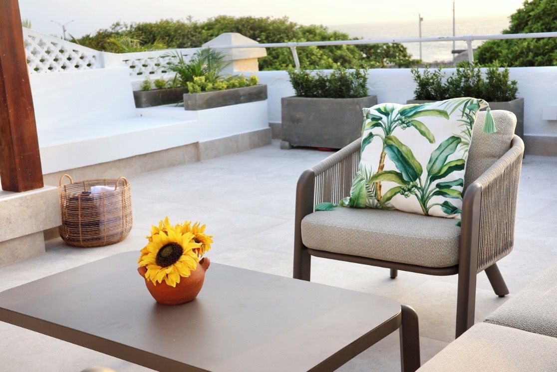 Un espacio al aire libre bañado por el sol con una silla de mimbre, una mesa con un jarrón de girasol y vistas al mar.