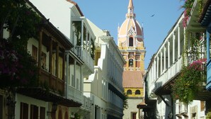 A narrow street with buildings on both sides, a prominent clock tower in the background, and people walking. - Colonial townhouse in the Old City with panoramic ocean views (Cartagena)