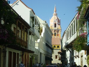 A narrow street with buildings on both sides, a prominent clock tower in the background, and people walking. - Colonial townhouse in the Old City with panoramic ocean views (Cartagena)