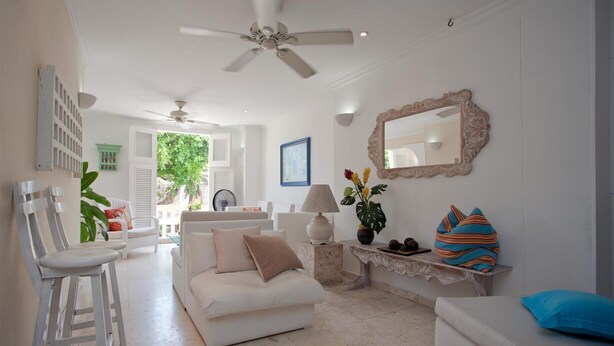 A spacious living room with a white sofa, a coffee table, and a ceiling fan. There is a large mirror on the wall and a window with shutters.