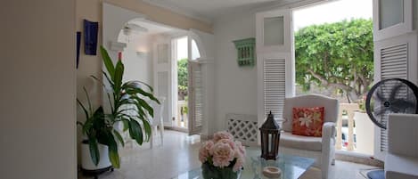 A spacious room with a glass coffee table, white furniture, and a view of greenery outside.