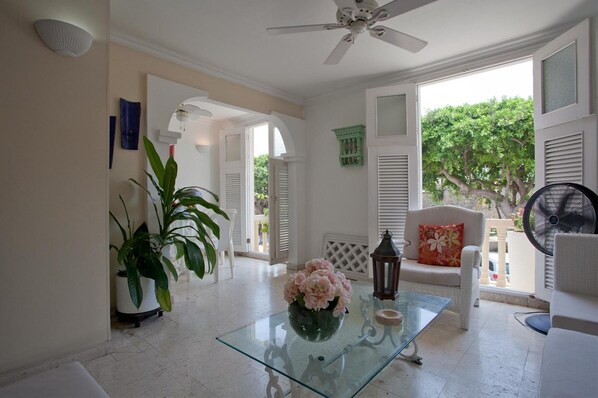 A spacious room with a glass coffee table, white furniture, and a view of greenery outside.