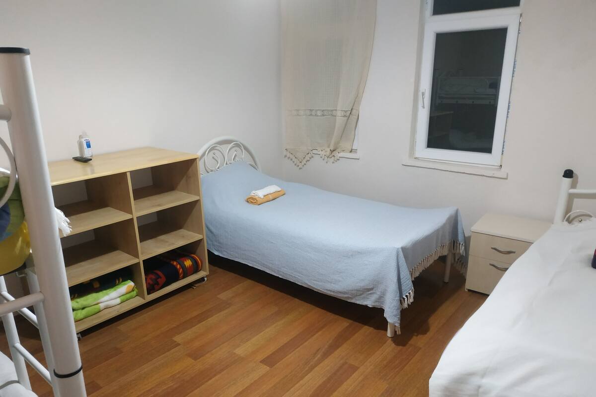 A small, tidy room with a single bed, a bedside table, a shelf unit, and a window.