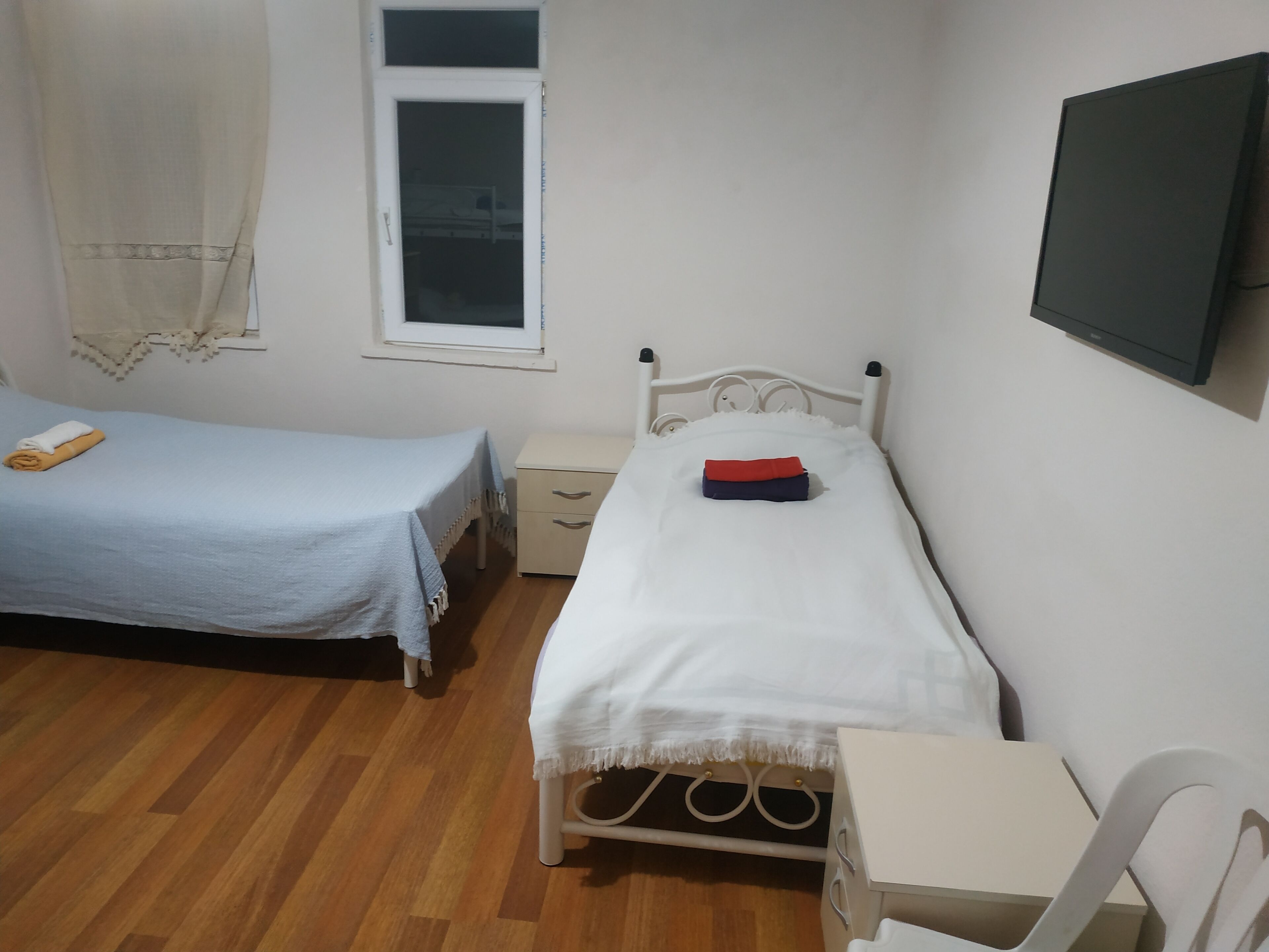 A small, clean room with two single beds, a bedside table, a chair, and a TV mounted on the wall.