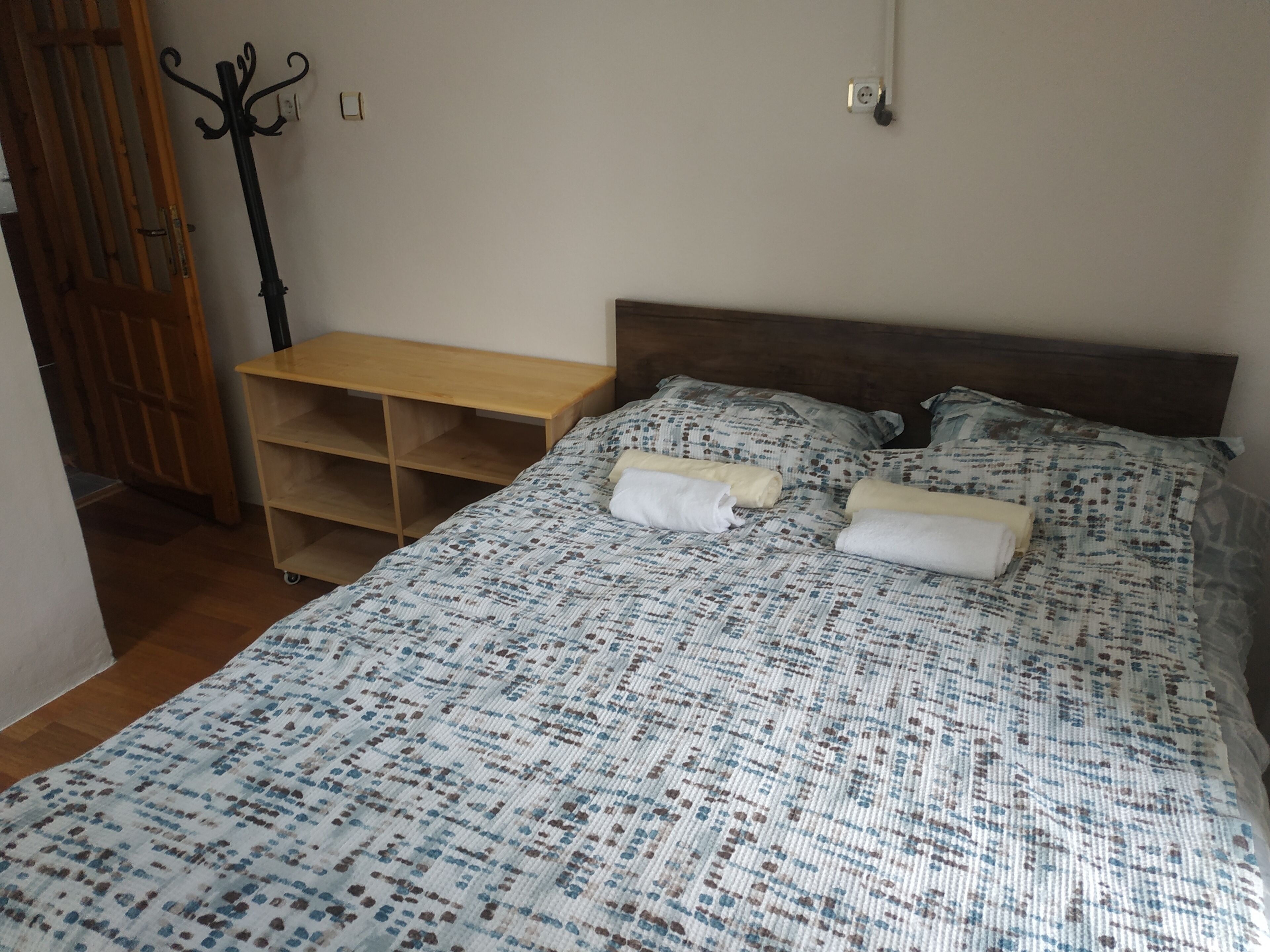 A bedroom with a bed, a wooden nightstand, a wall-mounted coat rack, and a door.